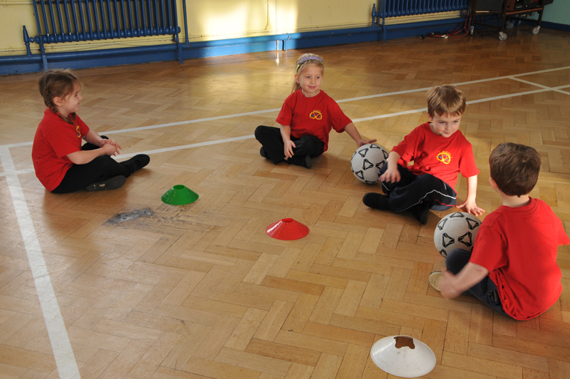 Four children in Manor Hill First School red shirts, sit on the gym floor with soccer balls and cones during a physical education class.