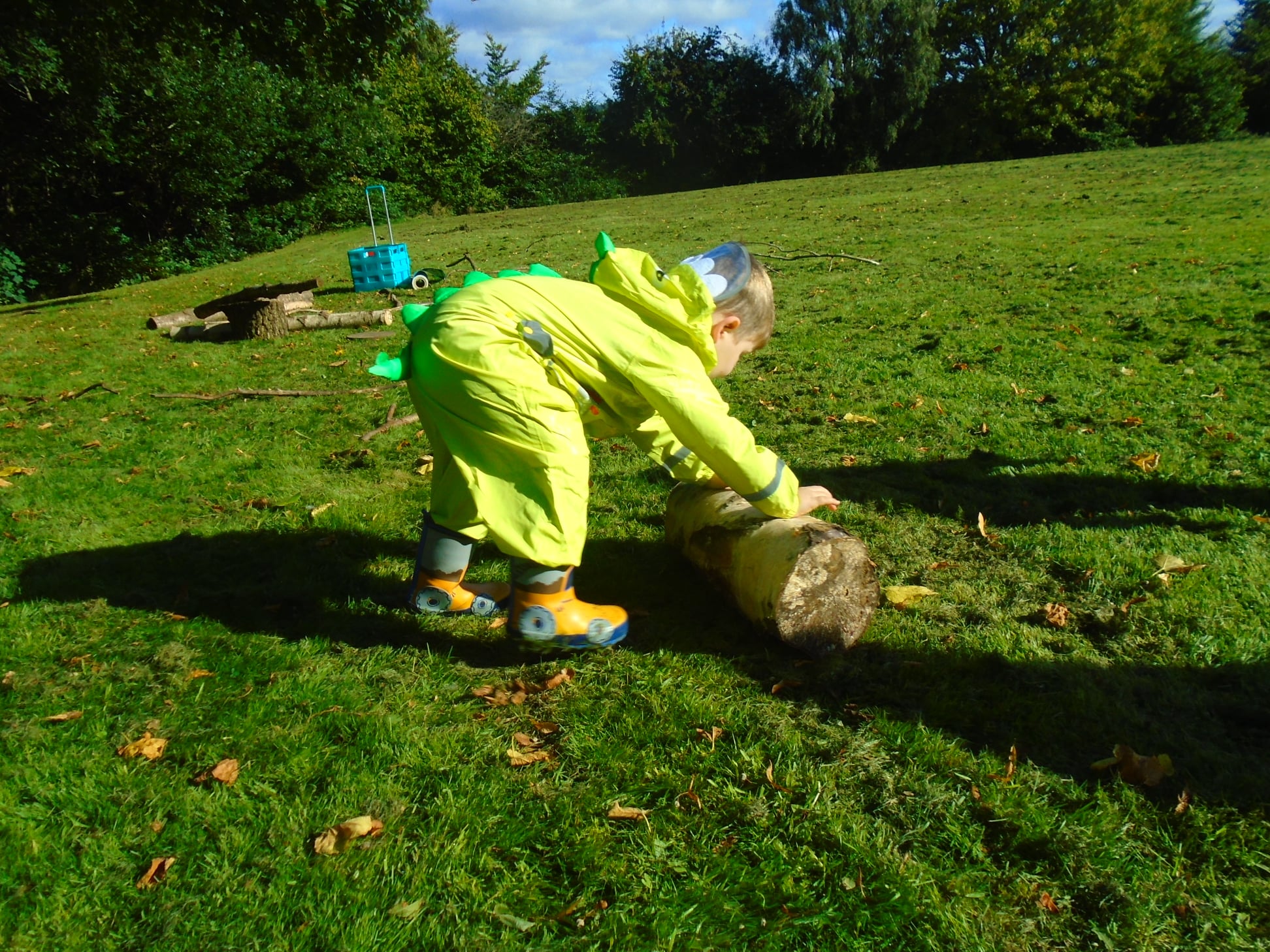 A child in a yellow rain suit plays with a log in a grassy field. This is Forest School, an activity encouraging exploration and curiosity in the natural world.