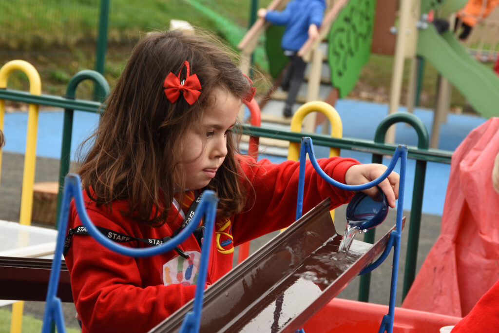 A Manor Hill First School nursery pupil plays outside on one of our playground sets. She is pouring water into a trough that runs along the set.